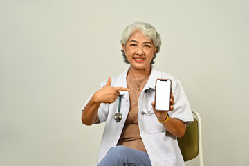 Confident senior female doctor showing smartphone with white blank screen for advertising medical mobile application or website