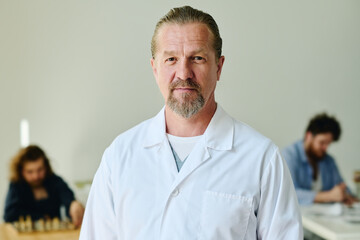 Confident mature psychiatrist in lab coat standing in front of camera against two patients of...