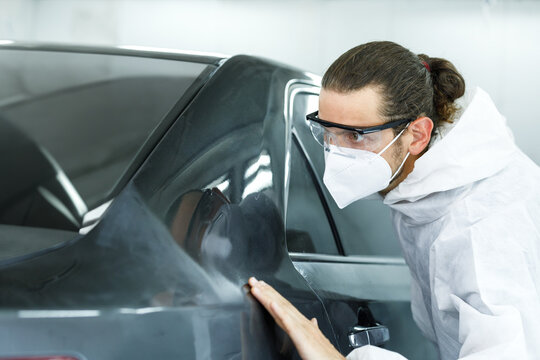 Young professional car painter wearing protective suit standing next to car in car painting room inspecting car body to look for scratches and damage