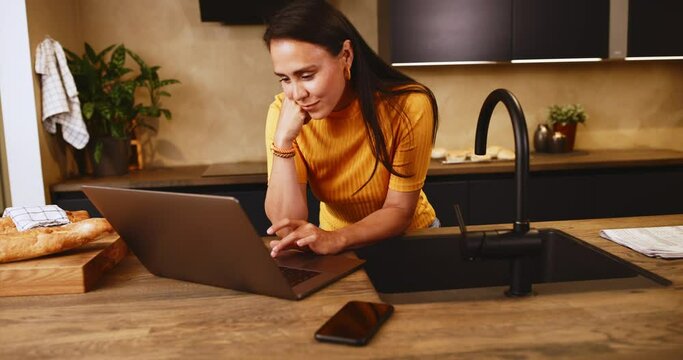 Smiling young woman working online with a laptop while leaning on a kitchen island at home