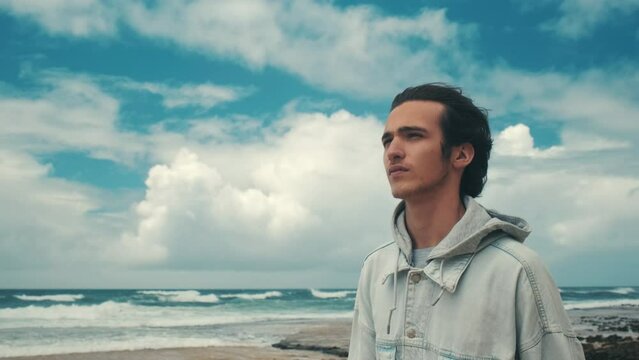 Young handsome man walks alone on the empty ocean beach on the cloudy day. Pretty guy walks a sandy shore and relaxing. Young Brunet man is walking against blue sea with clouds on the sky. Slow motion