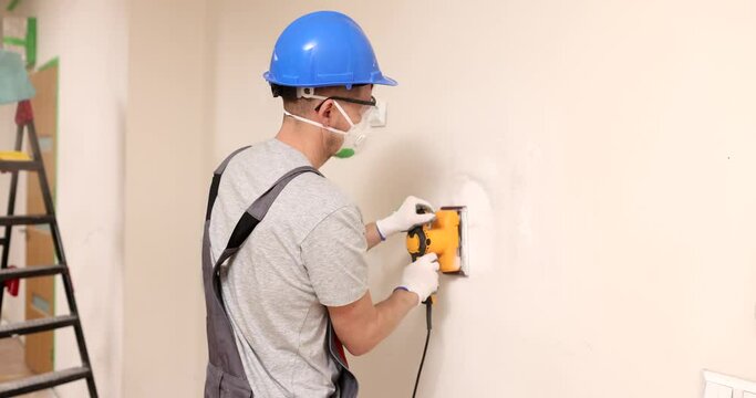 Home improvement worker in protective mask and goggles works with grinder to level surface of wall. Leveling walls with plaster