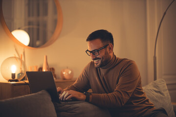 Man using a laptop, sitting on the sofa, enjoying himself at home, night time.