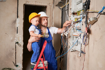 Male electrician in safety helmet holding child and pointing at electrical switchboard. Man and kid...