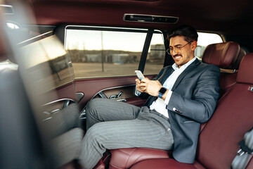 Young elegant businessman sitting on the back seat of a luxury car and using smartphone