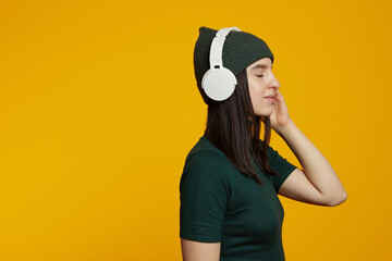 Side view of pretty young woman with white headphones listening relaxing music with closed eyes, isolated on yellow background with empty blank space