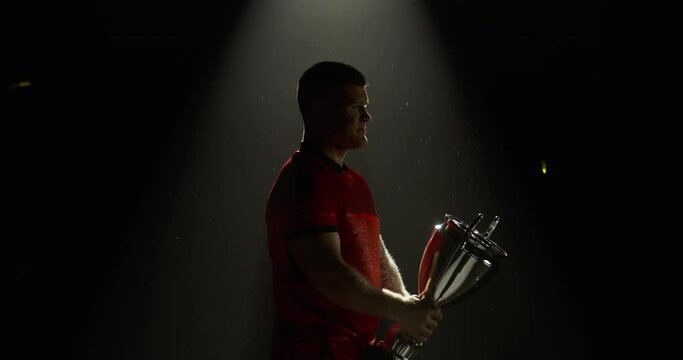 Silhouette of Caucasian male rugby player raising a trophy above head against bright light and falling confetti. Super slow motion, shot on RED cinema camera