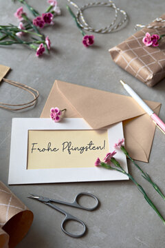 Text Frohe Phingsten means Happy Pentecost in German language. Envelope and greeting card with fresh carnation flowers. Wrapped gift, paper postcard, paper envelope, cord, and scissors on table.