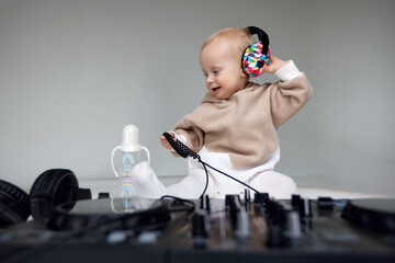 A cute smiling blonde baby in a beige hoodie sits on the floor and plays with dj headphones and a dj mixing board. Music and fun. Isolated on a gray background.