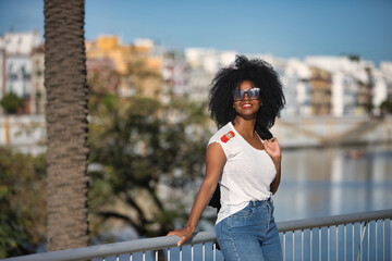 Young, beautiful, black woman with afro hair, white t-shirt and sunglasses leaning on a metal railing, smiling and happy receiving the sun's rays. Concept smile, happiness, success, current, modern.