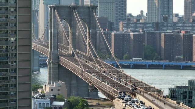 Aerial reveal of Brooklyn Bridge connecting Manhattan and Brooklyn, New York over the East River. American flag waving atop iconic suspended bridge in golden hour light.