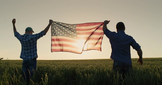Two men raise up the US flag against the backdrop of a wheat field at sunset