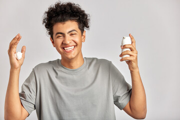 Young handsome smiling guy with black curly hair squeezing skincare cream from the dispenser to his hand. Biracial man in studio with healthy skin procedure. Wellness, cosmetics and skincare for men.