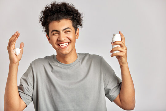 Young handsome smiling guy with black curly hair squeezing skincare cream from the dispenser to his hand. Biracial man in studio with healthy skin procedure. Wellness, cosmetics and skincare for men.