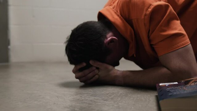 A prison inmate lays on ground crying, praying, weeping, sobbing next to bible while wearing orange prison uniform jumpsuit. Religious, Christian, Christianity.