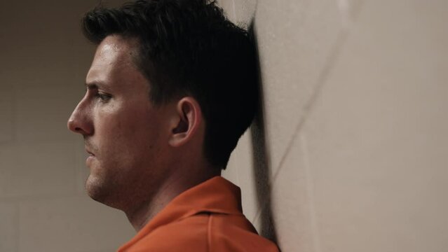 An unhappy man, sad, depressed attractive, handsome man, criminal prisoner, inmate in orange jumpsuit sitting in prison cell.