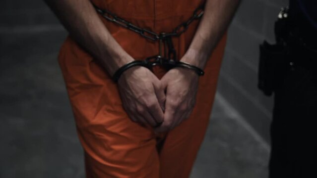 Hands of prison inmate in handcuffs, handcuffed in shackles, arrested for crime walking with prison guards to cell. A criminal prisoner, inmate in orange jumpsuit prison uniform.