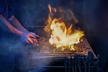 Hands of a blacksmith holding tongs with a workpiece in the blazing fire on the forge to get the...