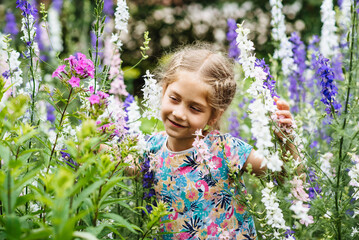 A funny little girl has fun in the summer outdoors. A carefree child runs and jumps among blooming summer flowers. Portrait of a beautiful girl smiling contentedly while smelling flowers in the garden