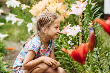 A little girl in the garden sniffs white lily flowers. Spring blooming concept. The child is having fun in the summer blooming garden of the garden. Childhood, harmony with nature, aromatherapy.