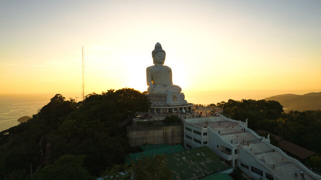 Big Buddha at sunset view from a drone. Phuket. People climb the steps to the statue. The green hills of the island are all around. In the distance, the bright sun goes over the sea. View from above