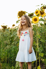 The cute little girl in a white dress with long beautiful hair laughing in a field of sunflowers. Portrait of a beautiful child enjoying nature at sunset. Little explorers, a way of life.