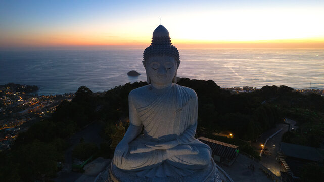 Big Buddha at sunset view from a drone. Phuket. People climb the steps to the statue. The green hills of the island are all around. In the distance, the bright sun goes over the sea. View from above