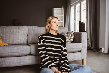 Obraz premium Young Woman Sitting on the Floor, Leaning Against a Sofa, Gazing Out of the Window