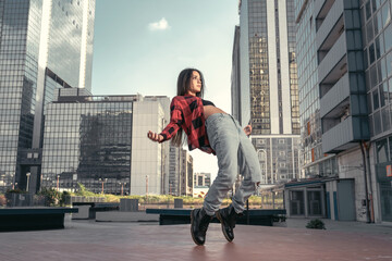 Photo shoot of stylish caucasian hip hop dancer posing on her toes with red shirt, jeans and black boots shoes and top with a urban town as background. City shooting of moving model bending the back