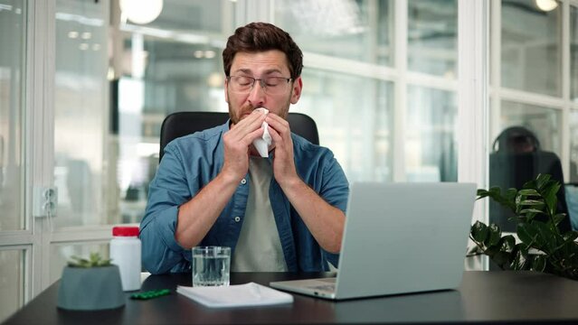 Ill, tired, sick businessman working in office sneezing and blowing nose with a tissue. Berded corporate male suffering from a cold, fever or flu while using a laptop at the workplace, trying to work.