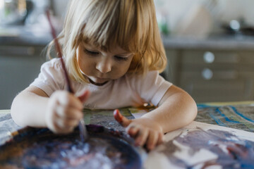 Cute little girl painting with tempera paint using a paintbrush. - obrazy, fototapety, plakaty