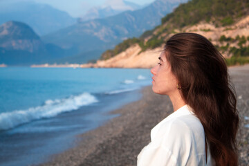 Fototapeta na wymiar a girl with long brown hair stands with her back, wearing a white shirt, against the background of mountains, sea and sand