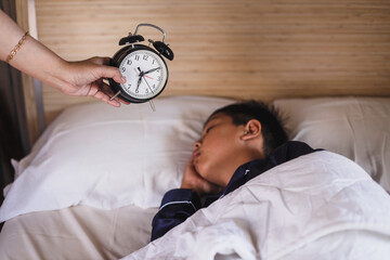 Hand of woman holding alarm clock and waking up her oversleep son at morning