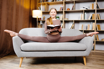 Young beautiful blonde woman in brown leggings and top sitting on a twine at home on the couch. The girl is reading a book sitting on the couch in an unusual position