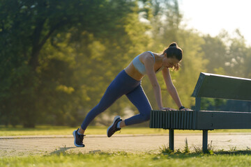 Full lenght portrait of beautiful fitness woman doing mountain clibers exercise against park bench.