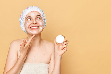 Spa wellness retreat. Skin care routine. Moisturizing after bath. Smiling woman wrapping in towel with shower cap on head cream in her hands isolated over beige background, copy space.