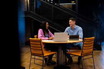 Happy diverse male and female colleague using tablet and laptop in casual office meeting