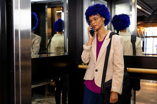 Happy biracial casual businesswoman with blue afro talking on smartphone in office elevator