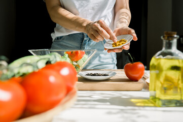 Crop anonymous woman in white shirt and jeans sprinkling healthy salad with spices while cooking vegetarian food in kitchen during sunny day