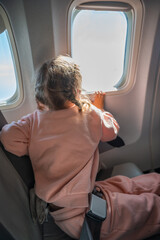 Cute little girl traveling by an airplane. Child sitting by aircraft window and looking outside. Traveling with kids abroad. 