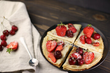 delicious fragrant pancakes folded in a triangle on a black plate and a wooden background and decorated with delicious ripe sweet strawberries, red fragrant cherries and coconut chips and honey