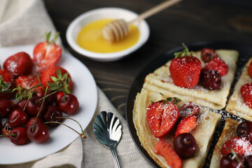 delicious fragrant pancakes folded in a triangle on a black plate and a wooden background and decorated with delicious ripe sweet strawberries, red fragrant cherries and coconut chips and honey