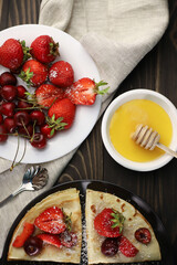 delicious fragrant pancakes folded in a triangle on a black plate and a wooden background and decorated with delicious ripe sweet strawberries, red fragrant cherries and coconut chips and honey
