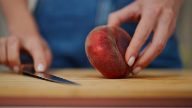 Close-up female hands halving flat peach in slow motion showing fruit. Closeup unrecognizable Caucasian lady cutting delicious ripe fruit with knife indoors