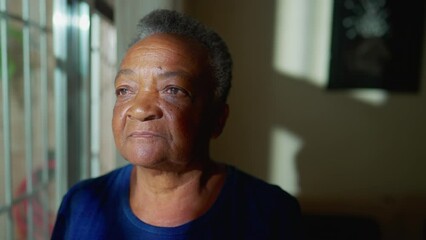 One contemplative black Brazilian senior woman standing by window looking out. A pensive African American elderly person in contemplation, close-up face - Powered by Adobe