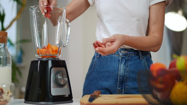 Young unrecognizable slim woman placing slices of flat peach in blender. Caucasian lady mixing ingredients for smoothie in kitchen at home