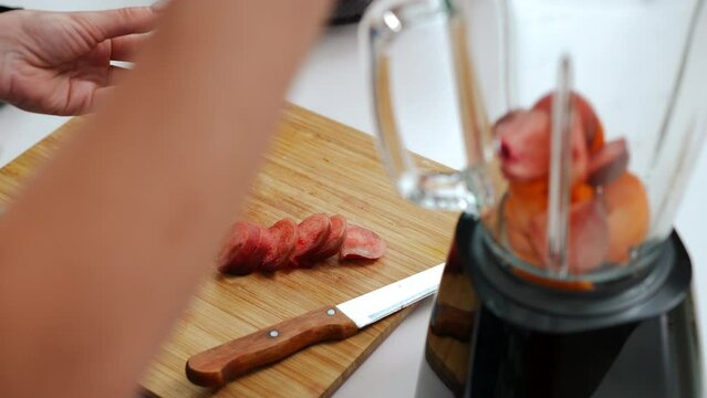 Close-up high angle view unrecognizable woman cutting flat peach placing slices in blender. Caucasian young housewife cooking tasty smoothie at home in kitchen