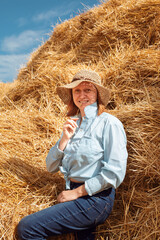 Naklejka na ściany i meble A woman in a shirt, jeans and a straw hat leans on a haystack and chews a spikelet