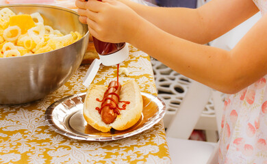 A little girl squeezing ketchup on a hotdog. Picnic, food, buffet outside. Child eating American...