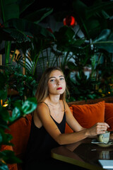 Young woman wearing black dress on thin straps siting on soft sofa at table in cafe among green plants inside arrangement. Thought Girl looking directly at camera. Hot drink cup on table. High quality